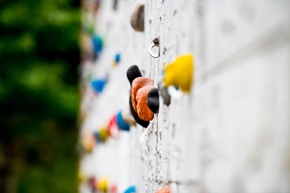 Nahaufnahme einer Kletterwand mit bunten Griffen und einem Sicherungshaken an einer grauen Wand.